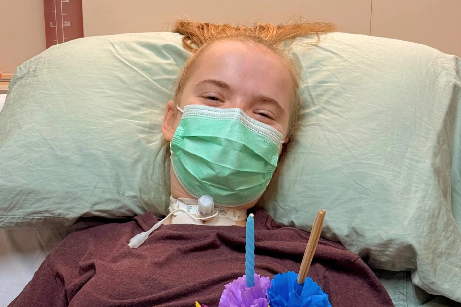 Makayla, a 16-year-old C3 complete quadriplegic patient, smiles from her hospital bed at Verita Neuro in Thailand, where she is receiving Epidural Stimulation treatment. She wears a green surgical mask and maroon top, with a visible tracheostomy at her neck. She holds colorful paper flowers and a candle from a celebration held during her recovery journey.